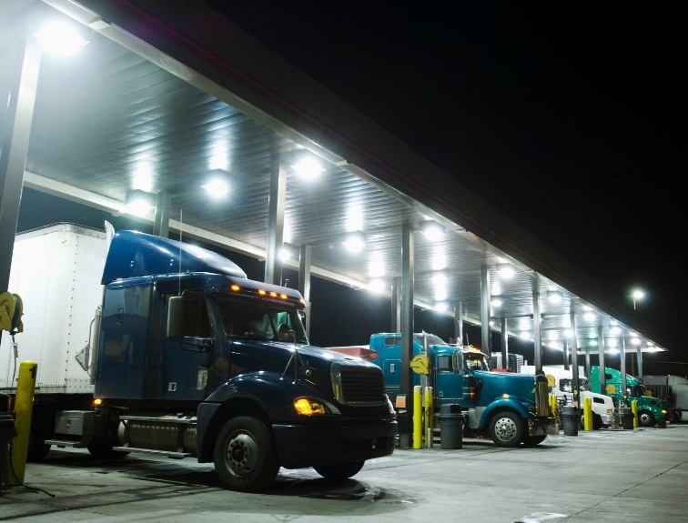 Semi-trucks parked at fuel islands under a brightly lit truck stop canopy at night.